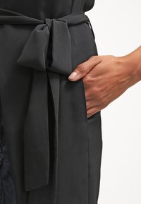 Hand mit neutralem Nagellack, die neben einem gebundenen Stoffgürtel auf einem schwarzen Kleidungsstück ruht, das die Textur und Passform hervorhebt.