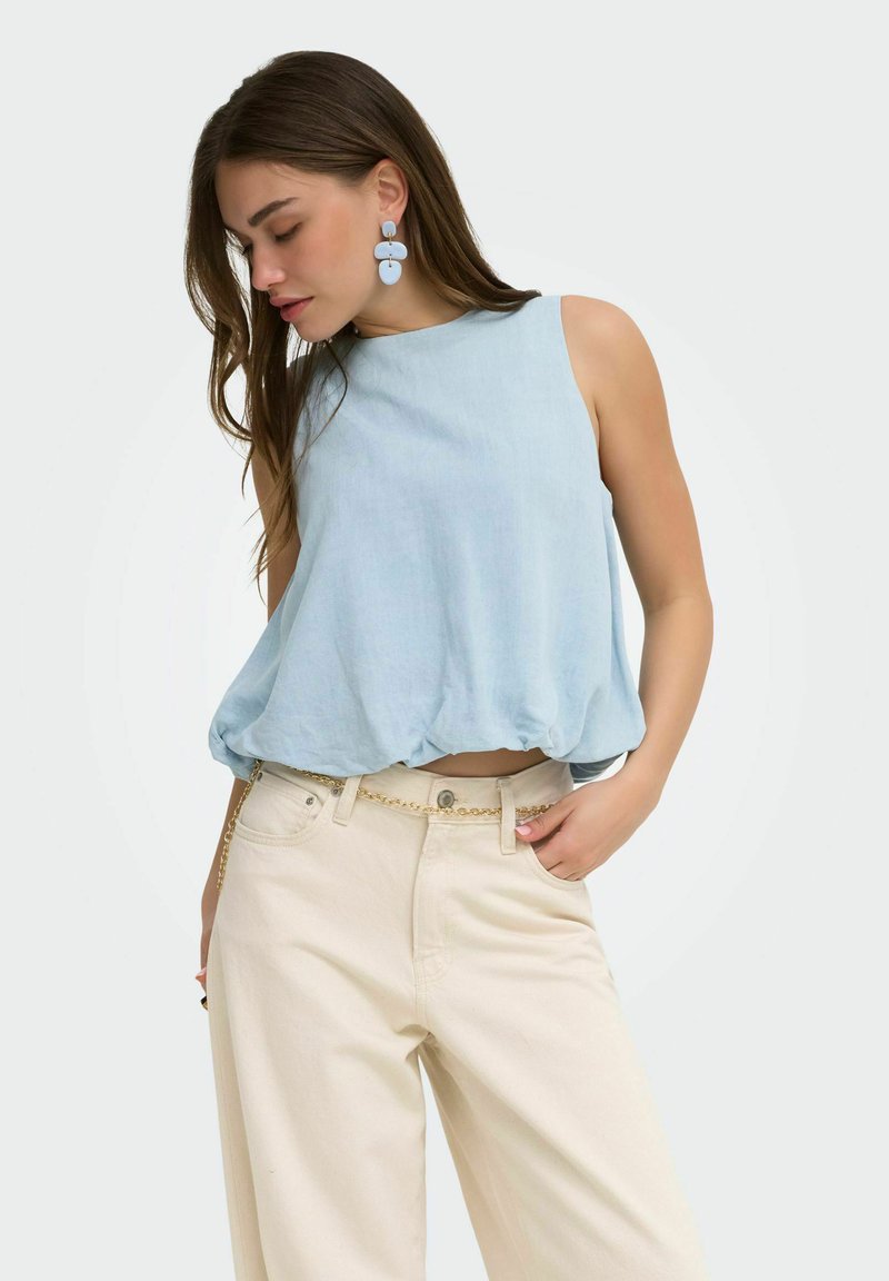 Femme portant un top sans manches bleu clair, un pantalon large crème avec une ceinture chaîne dorée, et de grandes boucles d'oreilles bleues pendantes, posant la tête baissée.