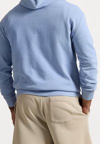 Person wearing light blue hoodie and beige pants with back pocket, hands in pants pockets, facing away from the camera.