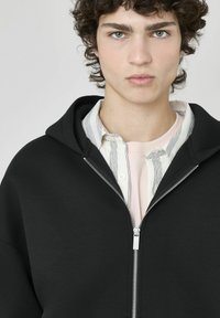 Haut à capuche zippé noir en tissu doux et texturé, avec une grande capuche et une fermeture éclair argentée, superposé sur une chemise rose clair à rayures.