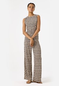 Mouwloze jumpsuit in beige en bruin geometrisch patroon, met een aansluitende top en wijde broekspijpen. Model staat met de handen gevouwen.