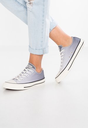 Osoba nosząca podwinięte, jasnoniebieskie podarte dżinsy oraz szare niskie trampki sznurowane z białymi podeszwami i czarnymi paskami.