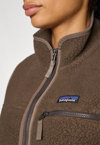Smeđa flis jakna s visokim ovratnikom, prednjim zipom i džepom na prsima. Sadrži Patagonia logo u plavoj i crnoj boji. Mekana tekstura.