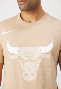 Homme portant un T-shirt beige avec un grand graphique blanc de tête de taureau et un petit logo blanc Nike sur la partie supérieure gauche de la poitrine.