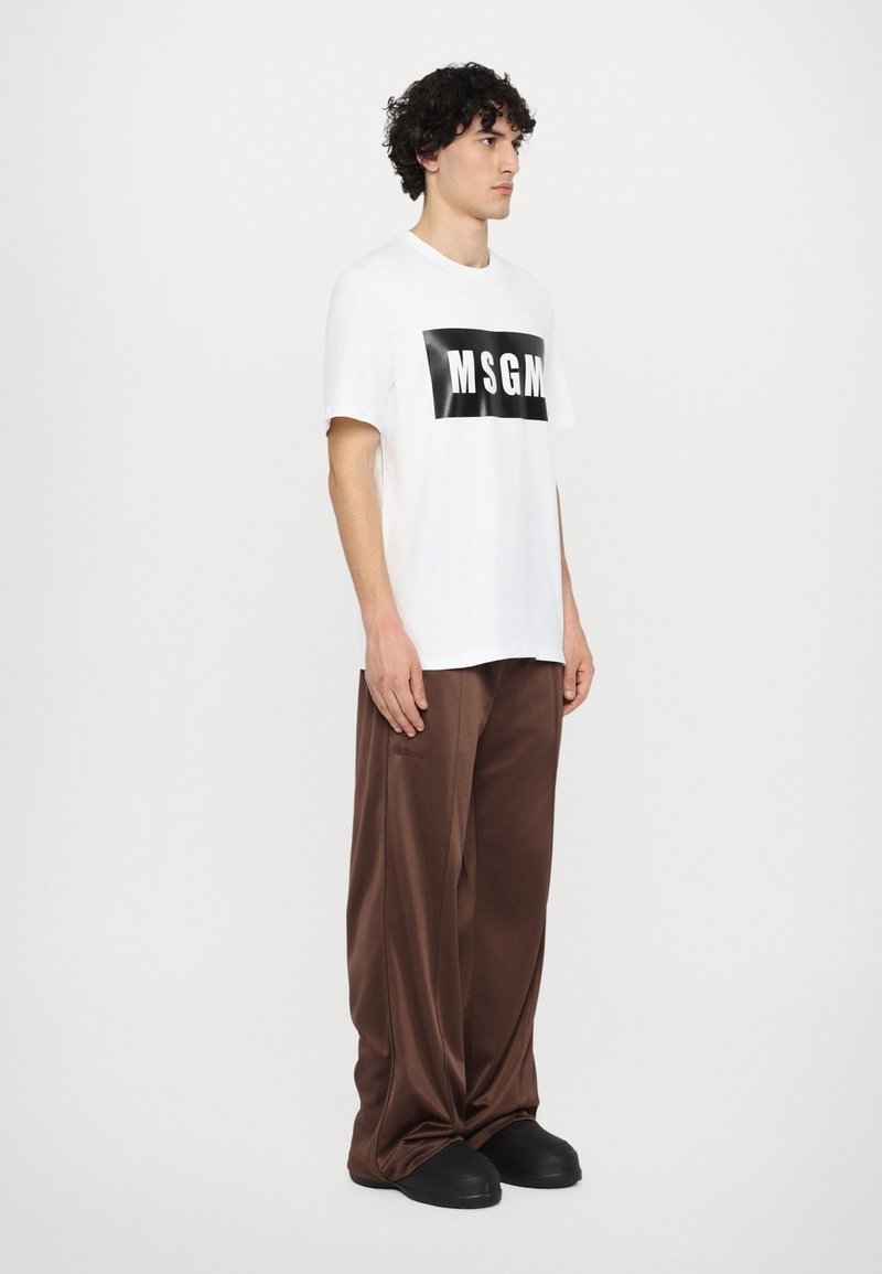 Hvid t-shirt med sort "MSGM" logo, parret med løstsiddende brune bukser og sorte slip-on sko med en glat tekstur.