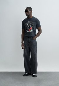 Άνδρας που φοράει μαύρα γυαλιά ηλίου, μαύρο γραφιστικό μπλουζάκι με πάνθηρα και κόκκινα λουλούδια, μαύρο τζιν με φαρδιά μπατζάκια, ασπρόμαυρα παπούτσια, όρθιος.