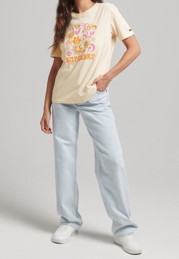 Camiseta amarilla clara con diseño floral en rosa y naranja, combinada con jeans rectos de pierna azul clara y zapatillas blancas.