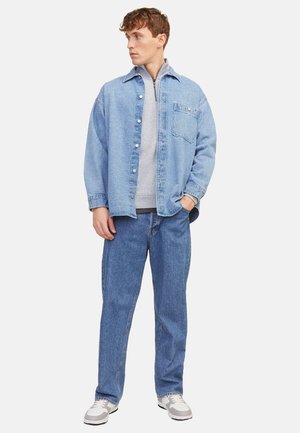 Nuori mies seisoo, yllä vaaleansininen farkkutakki, harmaa vetoketjullinen collegepaita, siniset farkut ja valkoiset lenkkarit, katsoo vasemmalle.