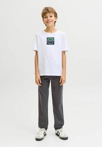 Un garçon souriant se tient debout, portant un t-shirt blanc avec un logo, un pantalon gris foncé et des baskets blanches, sur un fond clair uni.
