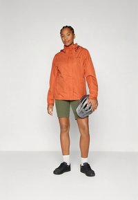 Oranžinė neperšlampama striukė su aukštu apykakliu, užtrauktuku priekyje ir reguliuojama kapišonu, dėvima virš žalių šortų. Laikomos juodos sportbačiai ir pilka šalmas.