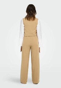 Jumpsuit beige ajusté avec jambes larges, buste sans manches, et chemise blanche à manches longues en dessous. Tissu lisse avec des lignes épurées.