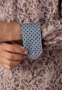 Camicia a maniche lunghe in tessuto floreale rosa, con un polsino blu decorato da motivi geometrici e un bottone visibile sul polso.