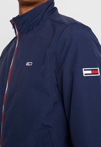 Tmavě modrá bunda s vysokým límcem, předním zipem, červeným a bílým kontrastním prošíváním a vyšitým logem na levém rukávu.