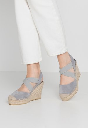 Feet wearing grey suede wedge espadrille sandals with cross straps paired with white cropped pants on a light background.