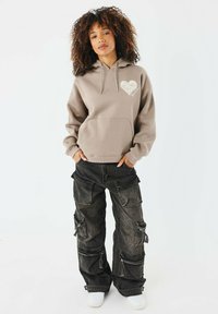 Sudadera con capucha marrón que presenta un gráfico de corazón en el pecho, combinada con pantalones cargo negros que tienen múltiples bolsillos y cremalleras.