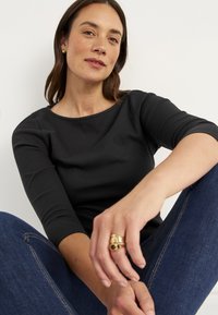 Schwarzes, geripptes Langarmshirt mit rundem Ausschnitt, getragen mit Denim. Die Hand des Models zeigt einen mehrteiligen Goldring. Der Hintergrund ist neutral.