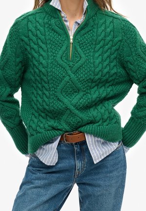 Iemand die een groene kabelgebreide trui met een ritssluiting van een kwart draagt over een blauw gestreept overhemd, gecombineerd met een blauwe spijkerbroek en een bruine riem.