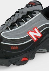 Μαύρο και γκρι αθλητικό παπούτσι New Balance 1000 με κόκκινες λεπτομέρειες, πλεκτό πάνω μέρος και χοντρή σόλα, αναδεικνύοντας το πλευρικό λογότυπο και τις λεπτομέρειες των κορδονιών.