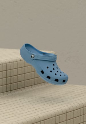 Un sabot bleu ajouré flotte au-dessus de marches carrelées beige, devant un mur uni.