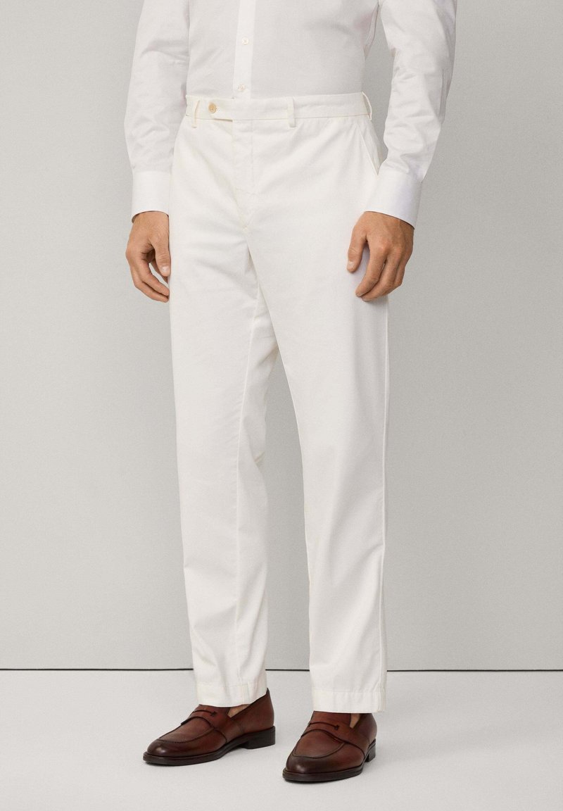 Pantaloni bianchi in cotone con taglio dritto, dotati di chiusura con un bottone e passanti per la cintura, abbinati a scarpe in pelle marrone.