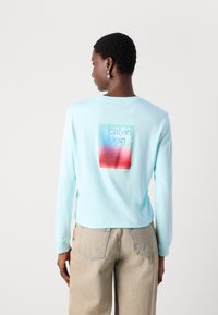 Hellblaues Langarm-T-Shirt mit einem quadratischen Grafikdesign, das auf dem Rücken den Text "calvin klein" in pastellfarbenen Farbverläufen zeigt.