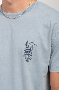 Camiseta de manga corta azul claro hecha de algodón suave, con un gráfico de grulla marina y hojas en el área del pecho izquierdo. Collar de cadena visible.