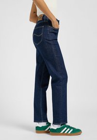 Jeans de mezclilla oscura con un corte recto, cinco bolsillos y costuras distintivas. Llevados con zapatillas verdes y blancas, que muestran un estilo casual.