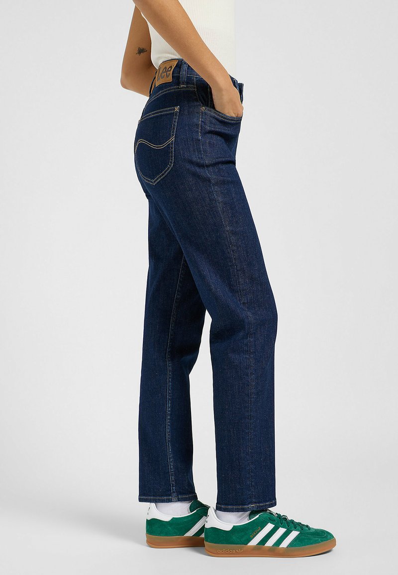 Granatowe dżinsy o prostym kroju, z pięcioma kieszeniami i charakterystycznym przeszyciem. Noszone z zielonymi i białymi sneakersami, co podkreśla swobodny styl.