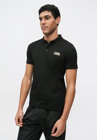 Fekete pamut poloing klasszikus gallérral. Bal oldalon a mellrésznél arany "KARL LAGERFELD" logóval díszített. Simított textúrájú, testhezálló szabású.