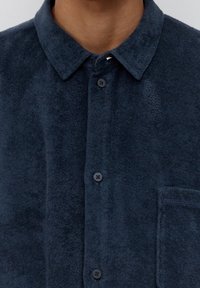 Mörkblå frottéskjorta med knäppning, mjuk textur, krage och bröstficka. Fyra knappar, minimal detaljering.
