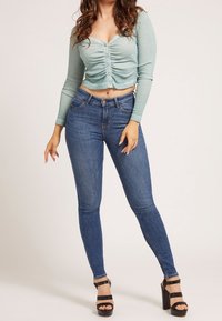 Žena v římské světle zelené vyhrnuté blůze, modrých skinny džínách s vysokým pasem a černých platformových podpatcích stojící na neutrálním pozadí.