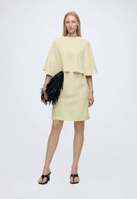 Femme portant une robe cape jaune clair et des sandales noires à talons hauts, tenant une grande pochette noire à franges.