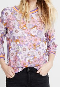 Vrouw in een paarse bloemige lange mouwen shirt met roze, witte en oranje bloemen en gestreepte paarse en oranje kraag en manchetten.