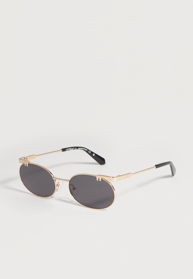 Lunettes de soleil ovales à monture dorée avec verres foncés et embouts de branches noirs posées sur une surface claire.