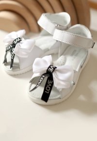 Sandali bianchi con una superficie sintetica liscia, cinturino regolabile e due fiocchi prominenti: uno bianco e uno nero con il testo 'LOVE'.