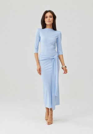 Žena v světle modrých dlouhých šatech s rukávy až na zem, s uzlem na boku, stojící, která má na sobě béžové sandály s otevřenou špičkou a zlaté náramky.