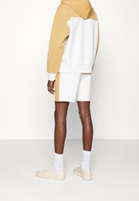 Huvtröja i beige och vit bomull med ett patchdesign, kombinerad med vita shorts. Beiga sneakers och vita strumpor fulländar looken.