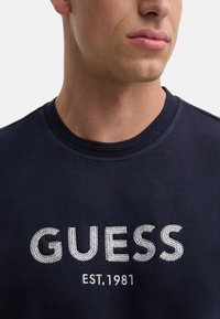 Felpa in cotone blu navy con scollo rotondo, caratterizzata da un logo "GUESS" ricamato in bianco e il dettaglio "EST. 1981" in cucitura argento.