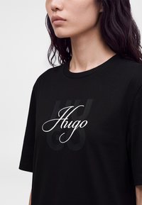 Γυναίκα με σκούρα μαλλιά φοράει μαύρο T-shirt με το λογότυπο "Hugo" σε λευκή γραμματοσειρά μπροστά, πάνω σε απλό φωτεινό φόντο.