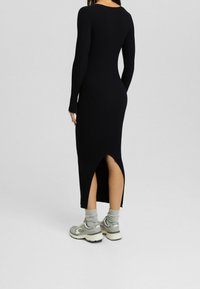 Femme portant une longue robe noire côtelée avec une fente au dos, des chaussettes grises et des baskets grises épaisses, debout devant un fond clair uni.