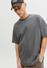 T-shirt gris à manches courtes en tissu mélange de coton avec une coupe décontractée et un col rond ; porté avec une casquette sombre ornée de texte blanc.