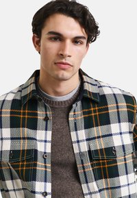 Koszula w kratę w kolorze zielonym, białym i pomarańczowym, z zapięciem na guziki, kołnierzem typu spread oraz przednimi kieszeniami. Sweter wełniany pod spodem.