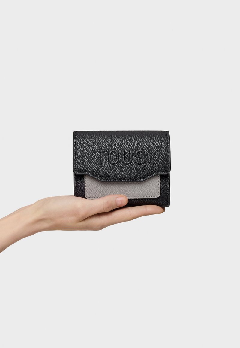 Black leather wallet with gray accent, embossed "TOUS" logo, flap closure, compact rectangular shape, held in a hand.