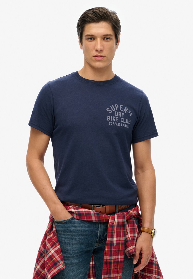 Marineblauwe katoenen T-shirt met korte mouwen en een ronde hals, met een grijs gedrukt logo op de linker bovenkant. Draagt een spijkerbroek en een rode geruite blouse om de taille geknoopt.