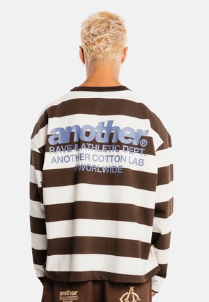 Persona con cabello rubio corto de espaldas que lleva una camisa de rayas marrón y blanca con texto grande en la parte trasera.
