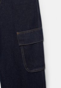 Mørkeblåt denimstof med en rektangulær cargolomme med klap og synlige orange sømme på siden af et beklædningsstykke.