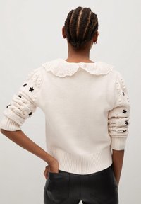 Pull crème avec un col en dentelle, des manches texturées avec des broderies florales noires, une taille en côtes et une silhouette ajustée.