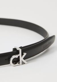 Juodas odinis diržas su sidabriniu "ck" logotipo sagtimi, lygiu paviršiumi ir įspaustu "Calvin Klein" prekės ženklu per visą diržo ilgį.