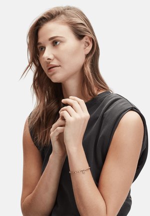 Roze gouden armband met een ronde charme en kraalaccenten, gedragen op een pols. Het model heeft lang, golvend haar en draagt een donker mouwloos top.