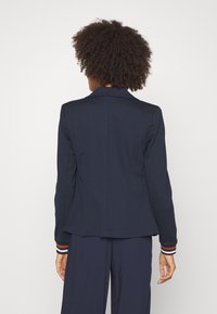 Blazer marine à la coupe structurée, doté d'un col cranté et de poignets rayés avec des accents rouges et blancs. Texture de tissu lisse.